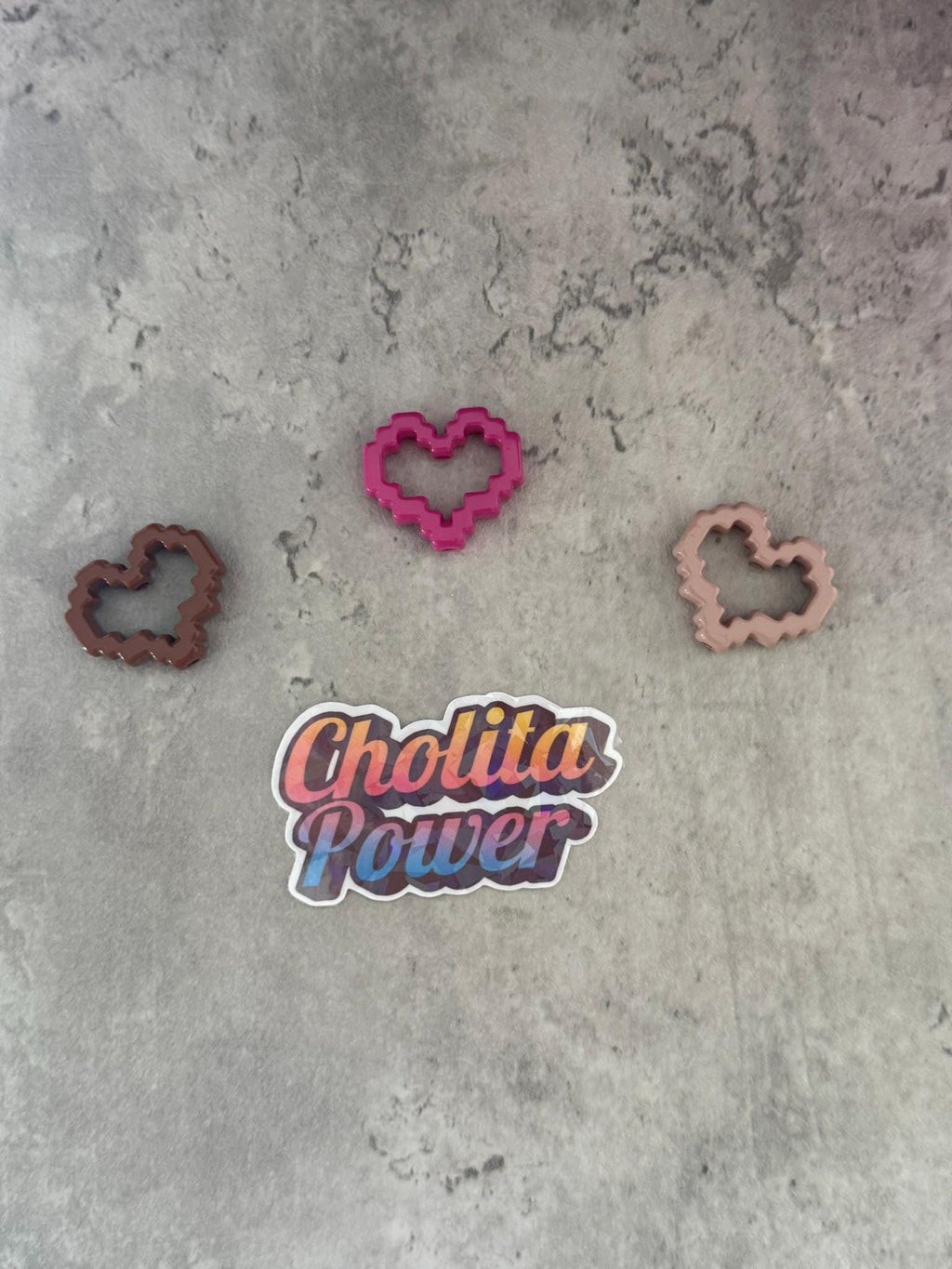 May include: Three pink, brown, and peach heart-shaped cookie cutters on a gray textured surface. A sticker with the text 'Chollita Power' is in the center of the image.