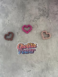 May include: Three pink, brown, and peach heart-shaped cookie cutters on a gray textured surface. A sticker with the text 'Chollita Power' is in the center of the image.