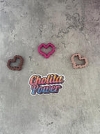May include: Three pink, brown, and peach heart-shaped cookie cutters on a gray textured surface. A sticker with the text 'Chollita Power' is in the center of the image.