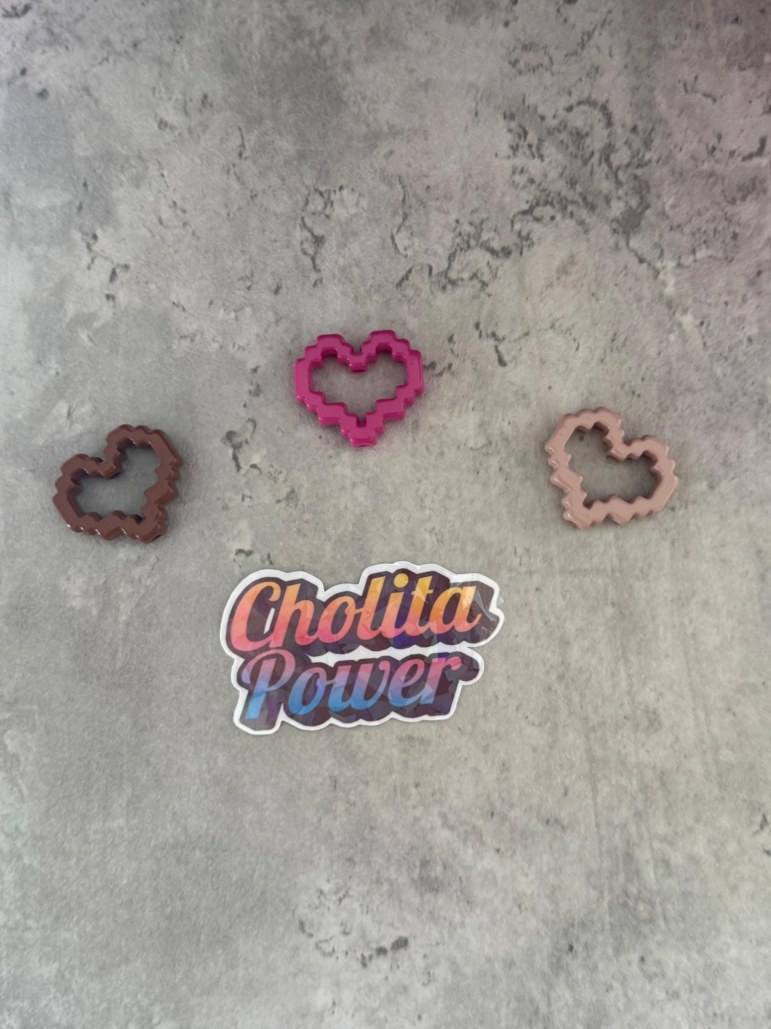 May include: Three pink, brown, and peach heart-shaped cookie cutters on a gray textured surface. A sticker with the text 'Chollita Power' is in the center of the image.