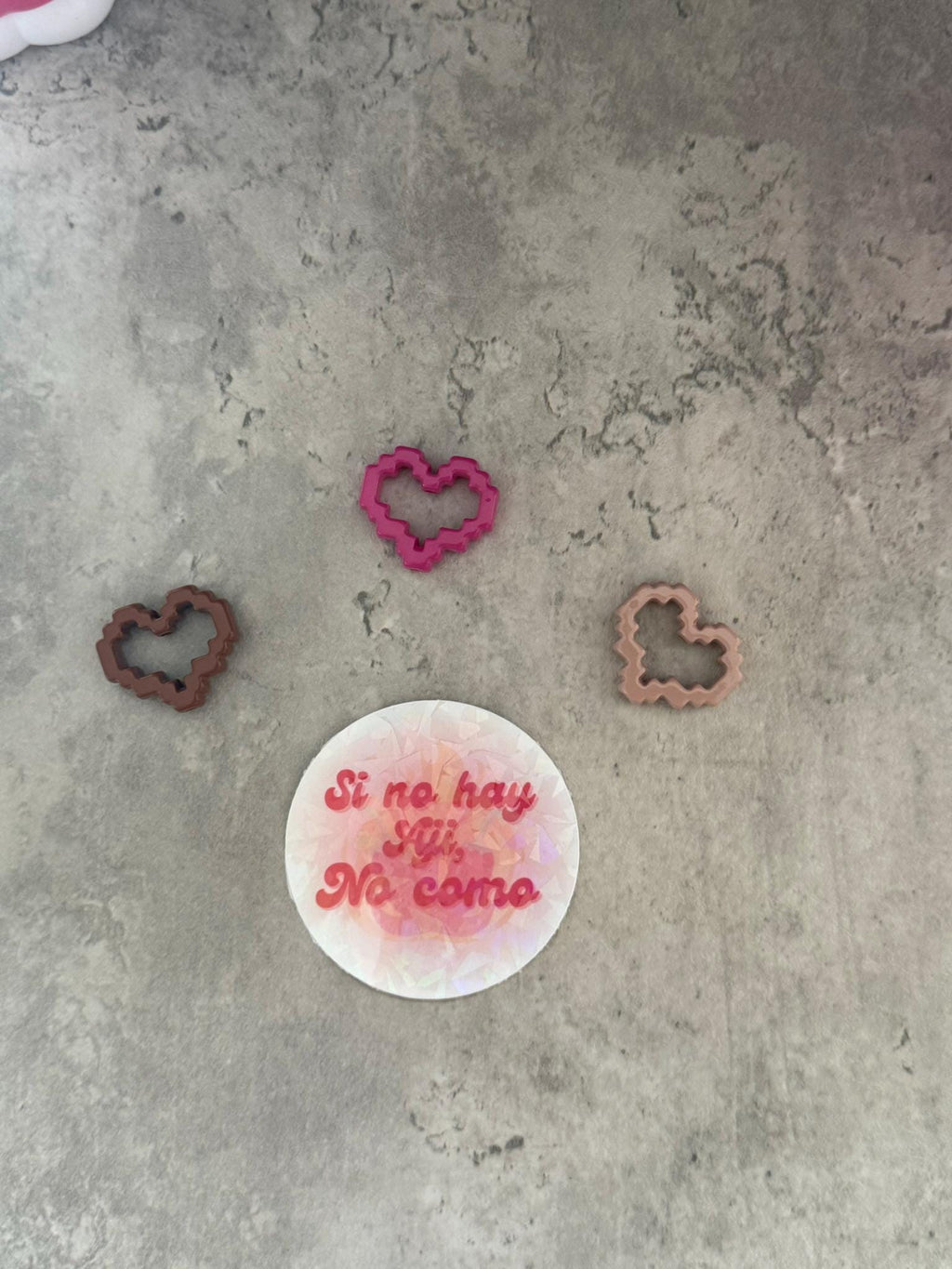 May include: Three heart-shaped cookie cutters, one pink, one brown, and one light brown. The cookie cutters are on a gray textured surface. A white round cookie cutter with the text 'Si no hay yji, No como' in pink is also on the surface.