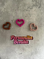 May include: Three heart-shaped cookie cutters, one pink, one brown, and one beige, on a gray textured surface. A sticker with the text 'Peru anita Bonita' is below the cookie cutters.