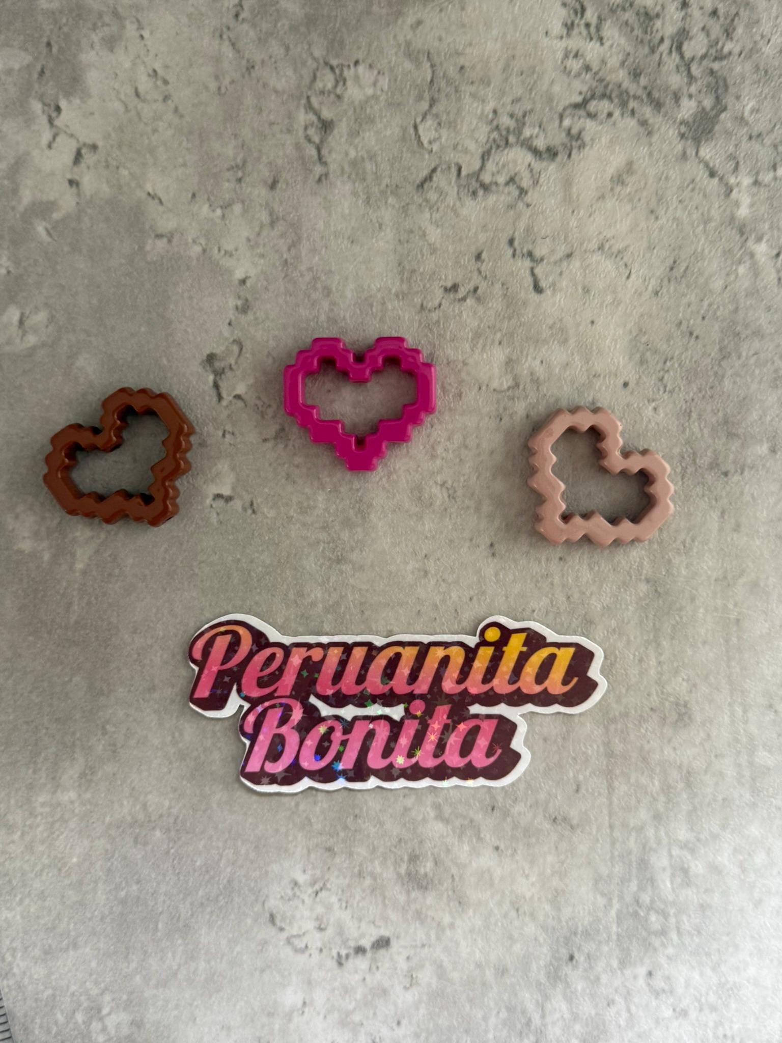 May include: Three heart-shaped cookie cutters, one pink, one brown, and one beige, on a gray textured surface. A sticker with the text 'Peru anita Bonita' is below the cookie cutters.