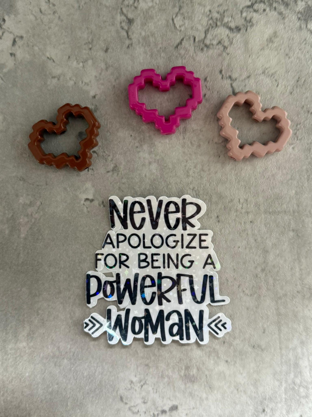 May include: A white sticker with black text that reads 'Never apologize for being a powerful woman' with two arrows pointing outwards on either side of the word 'woman'. The sticker is on a gray textured background with three hearts in the background. One heart is pink, one is brown, and one is light brown.