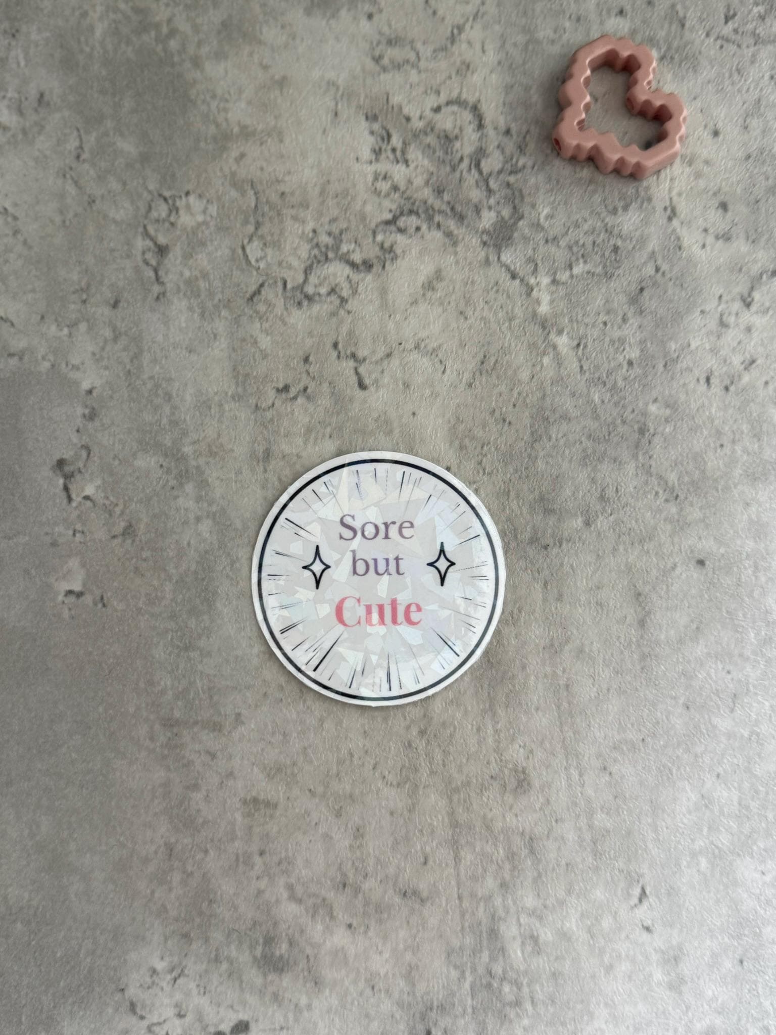 May include: A white circle sticker with a black border and black clock hands. The sticker has the text 'Sore but Cute' in pink and black text with two black diamond shapes on either side of the text.