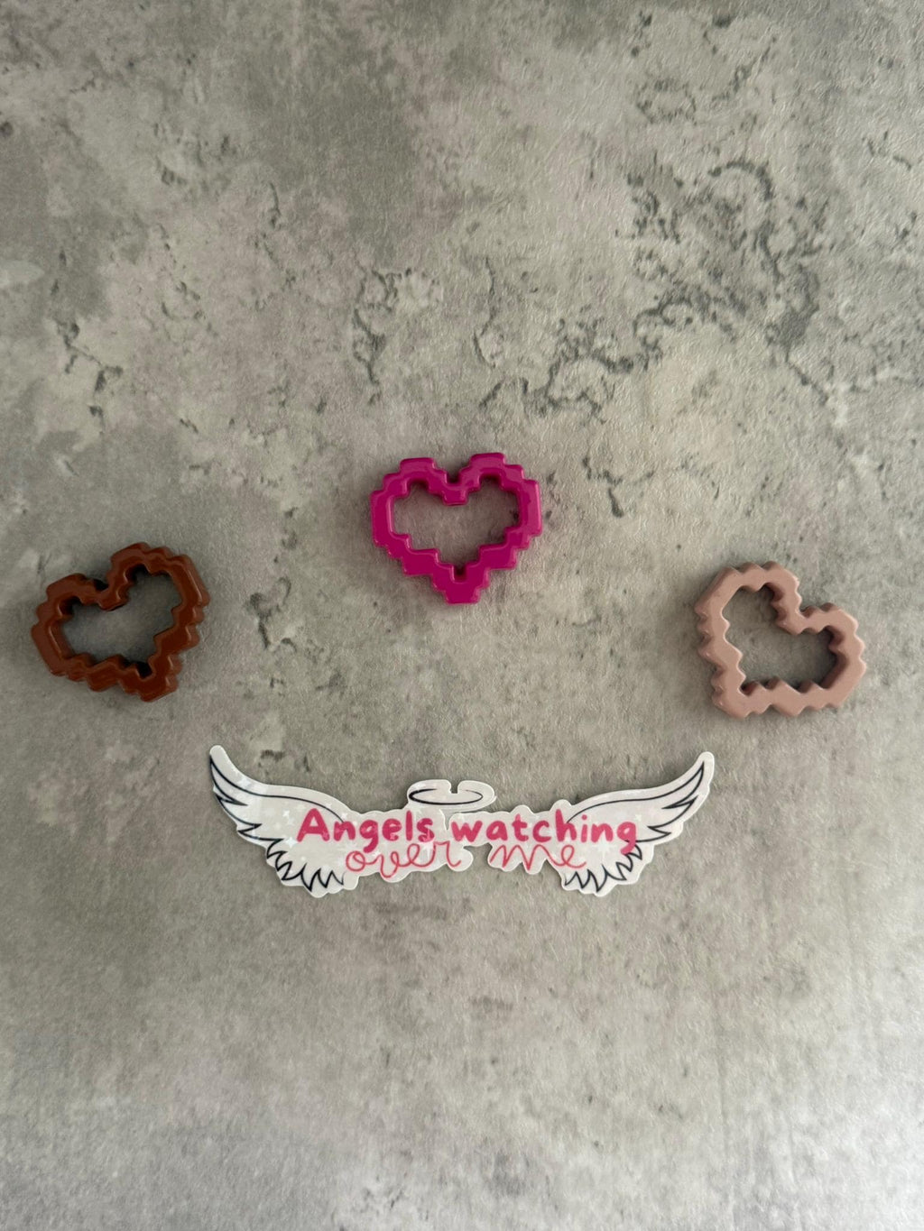 May include: Three pink, brown, and brown heart-shaped cookie cutters on a gray textured surface. The pink cookie cutter is in the center of the image. Below the cookie cutters is a white sticker with black wings and the text 'Angels watching over me' in pink.