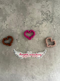 May include: Three pink, brown, and brown heart-shaped cookie cutters on a gray textured surface. The pink cookie cutter is in the center of the image. Below the cookie cutters is a white sticker with black wings and the text 'Angels watching over me' in pink.