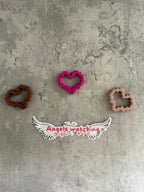 May include: Three pink, brown, and brown heart-shaped cookie cutters on a gray textured surface. The pink cookie cutter is in the center of the image. Below the cookie cutters is a white sticker with black wings and the text 'Angels watching over me' in pink.