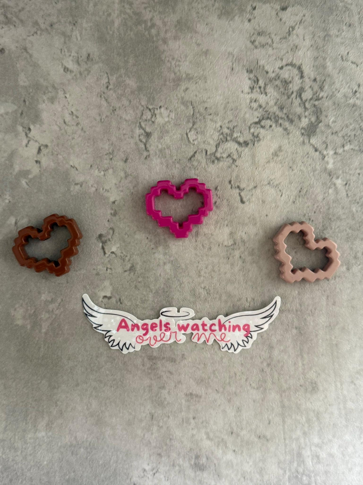 May include: Three pink, brown, and brown heart-shaped cookie cutters on a gray textured surface. The pink cookie cutter is in the center of the image. Below the cookie cutters is a white sticker with black wings and the text 'Angels watching over me' in pink.