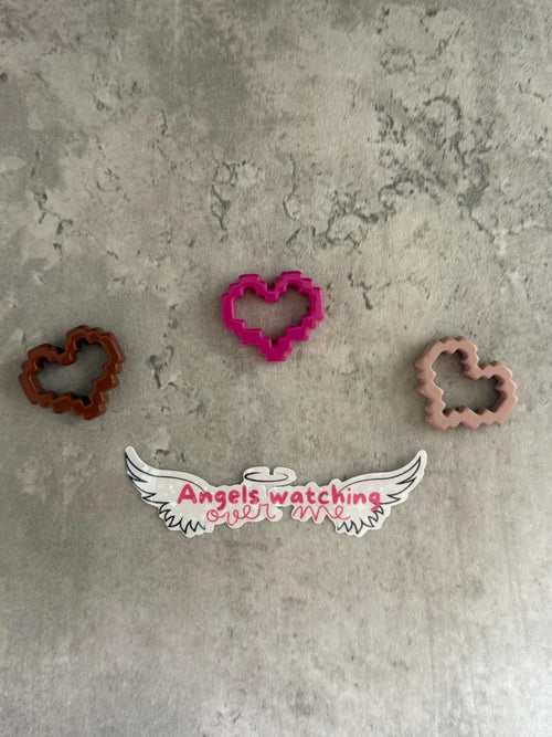 May include: Three pink, brown, and brown heart-shaped cookie cutters on a gray textured surface. The pink cookie cutter is in the center of the image. Below the cookie cutters is a white sticker with black wings and the text 'Angels watching over me' in pink.