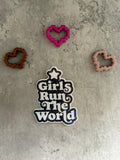 May include: A black and white sticker with the text 'Girls Run The World' in a retro font. The sticker has a star at the top and is surrounded by glitter. There are three hearts in the background, one pink, one brown, and one peach.