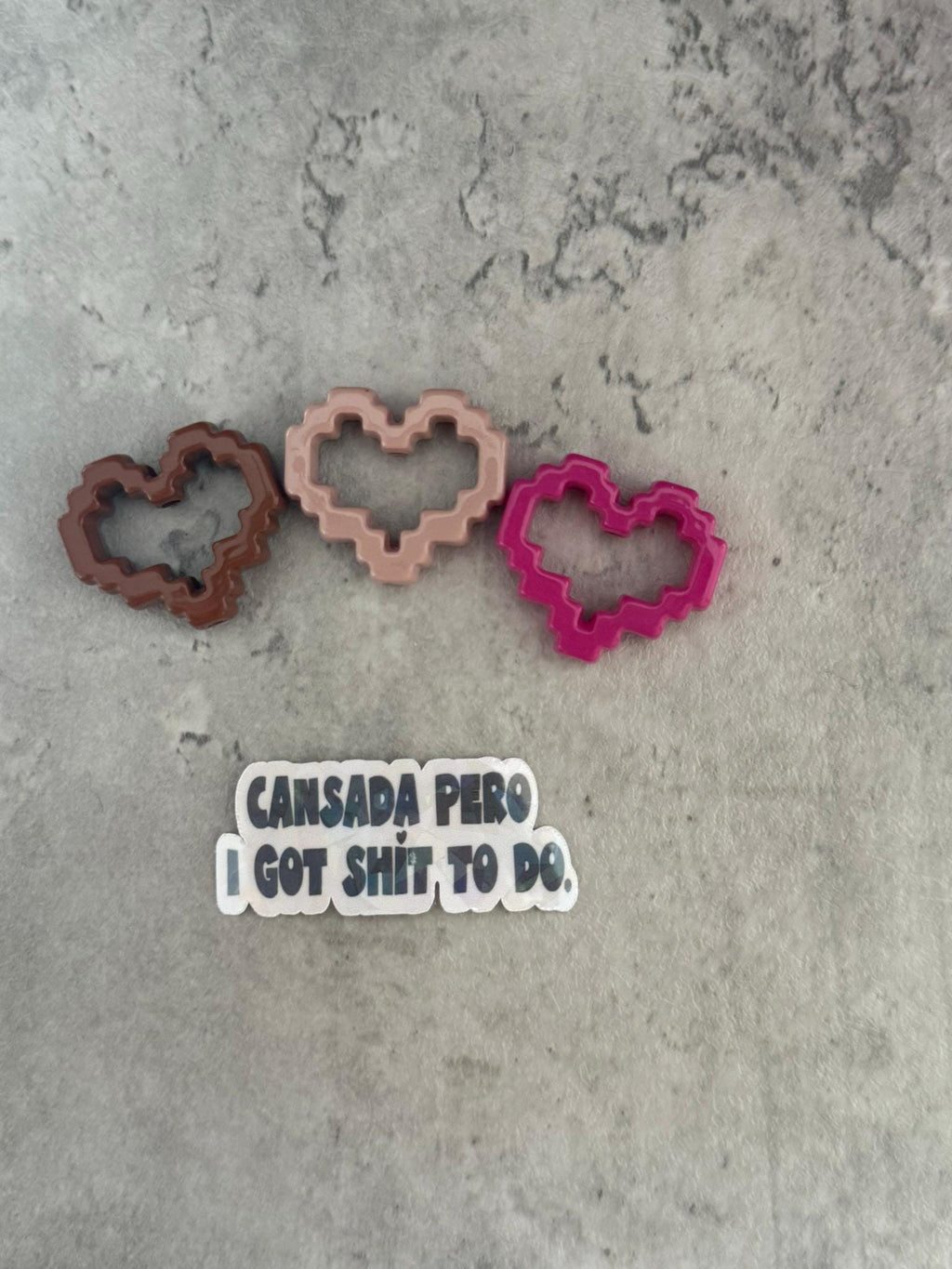 May include: Three pixelated heart-shaped cookie cutters in brown, pink, and red. The cookie cutters are on a gray textured surface. A sticker with the text 'Cansada Pero I Got Shit To Do' is below the cookie cutters.