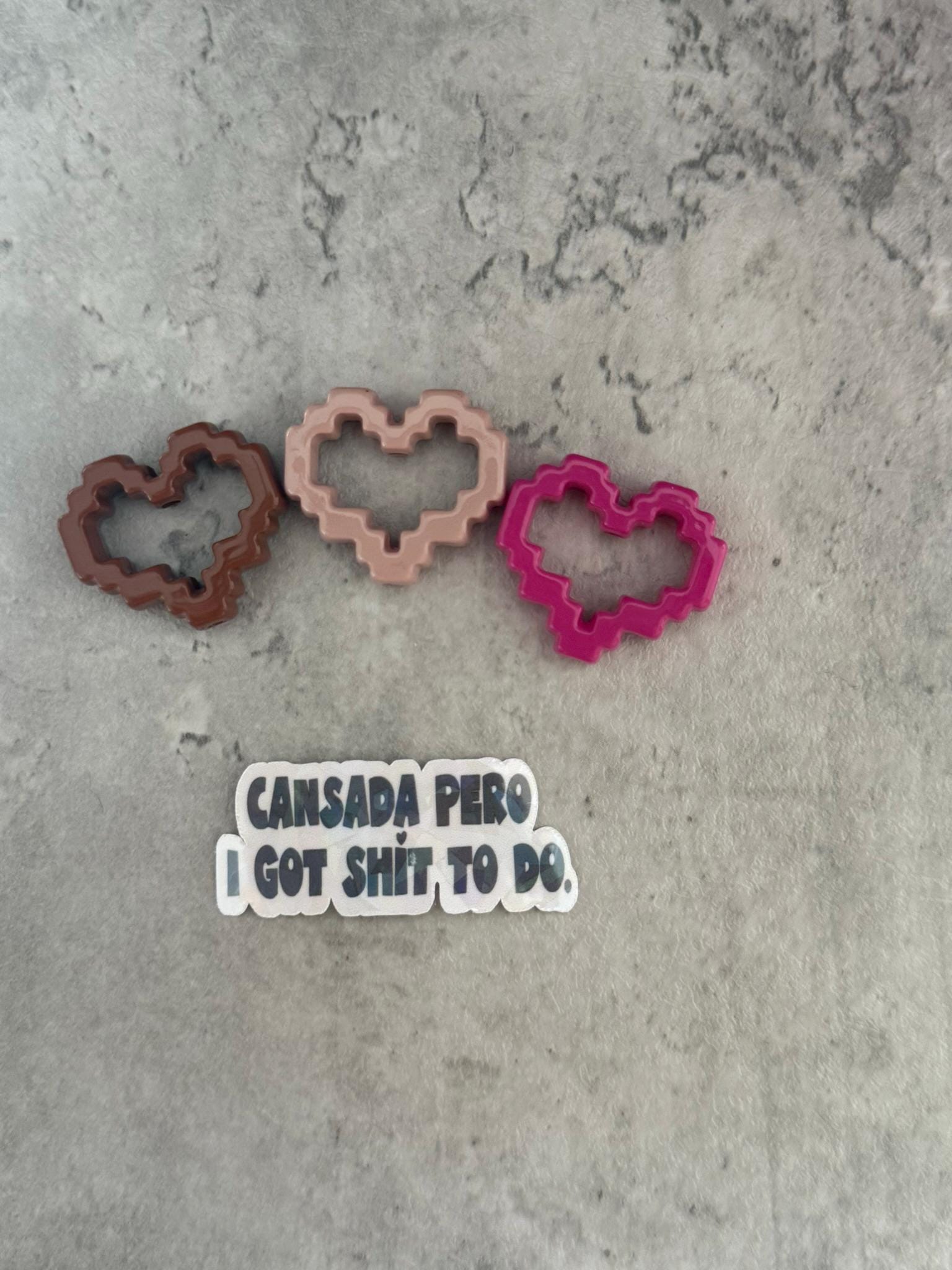 May include: Three pixelated heart-shaped cookie cutters in brown, pink, and red. The cookie cutters are on a gray textured surface. A sticker with the text 'Cansada Pero I Got Shit To Do' is below the cookie cutters.