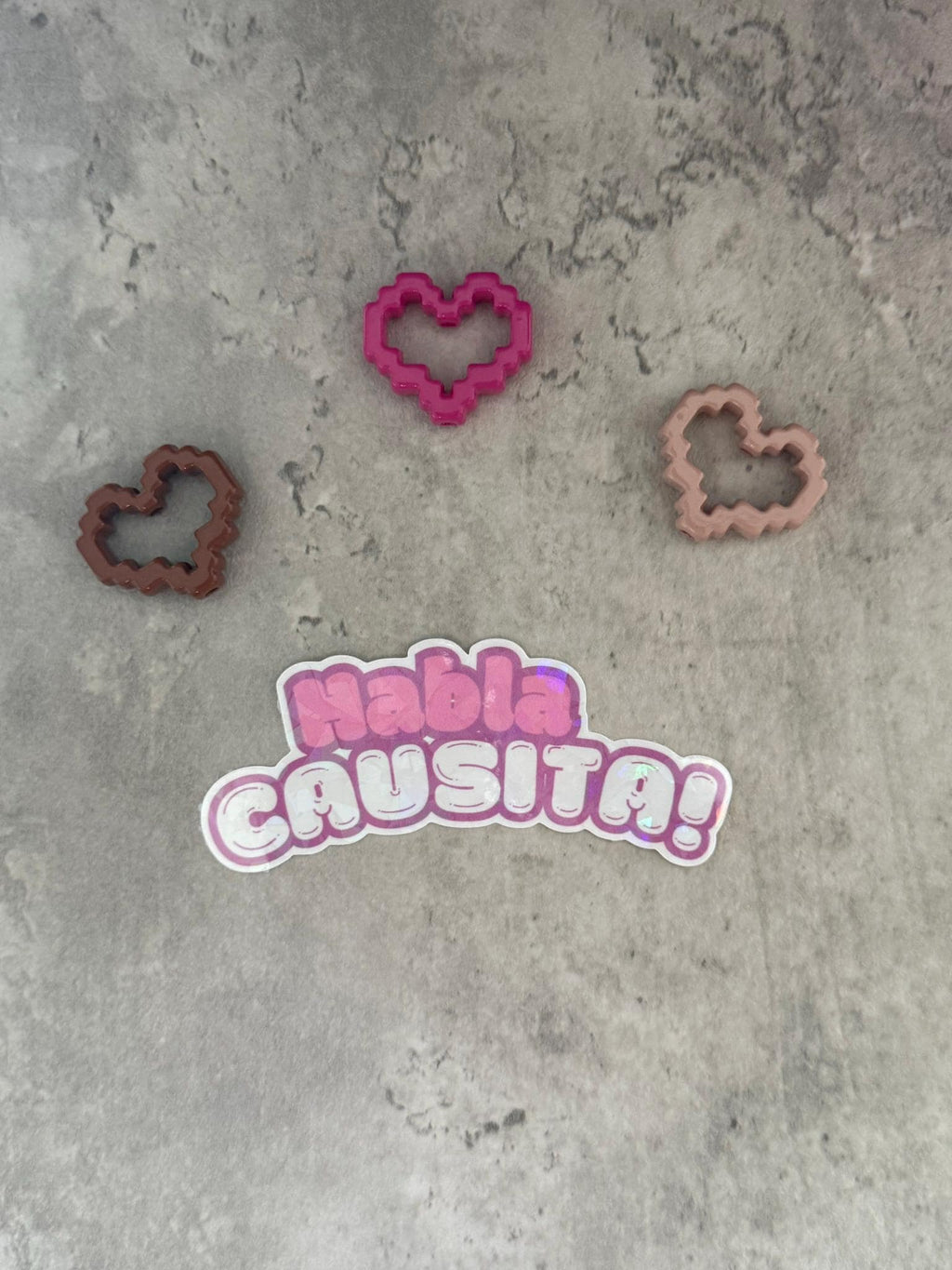 May include: A pink sticker with the text 'Habla Causita!' in a white outline. Three heart-shaped cookie cutters are on a gray background. One cookie cutter is brown, one is pink, and one is light brown.