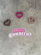 May include: A pink sticker with the text 'Habla Causita!' in a white outline. Three heart-shaped cookie cutters are on a gray background. One cookie cutter is brown, one is pink, and one is light brown.